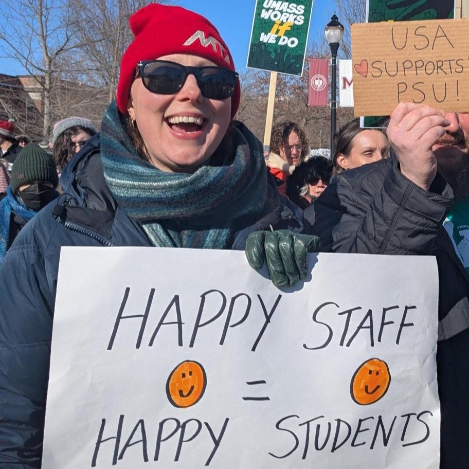 White woman in red knit had and sunglasses holding sign reading "Happy staff = happy students"